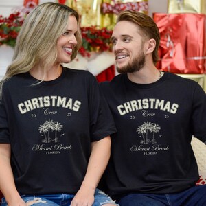 May include: A black short-sleeved t-shirt featuring the text CHRISTMAS CREW 20 25, an illustration of three palm trees, and the text Miami Beach FLORIDA in a light-colored font. The design is centered on the chest of the garment.