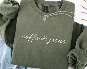 Puede incluir: Una sudadera verde oliva con las palabras "coffee & jesus" en escritura blanca. Un collar plateado con un colgante circular está drapeado en la parte delantera. La sudadera está doblada, con unos vaqueros azules visibles.