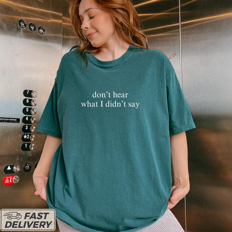 May include: Teal-colored t-shirt with the text "don't hear what I didn't say" in white. The shirt has a relaxed fit and short sleeves. The model is wearing the shirt in front of an elevator.