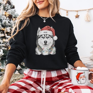 May include: Black sweatshirt featuring a Siberian Husky wearing a Santa hat with the name "Max" and a string of Christmas lights. The dog has gray, black, and white fur. The sweatshirt is a festive holiday design.