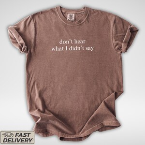 May include: A brown t-shirt with the phrase "don't hear what I didn't say" printed in white. The shirt has a crew neck and short sleeves. The fabric appears soft and slightly wrinkled.