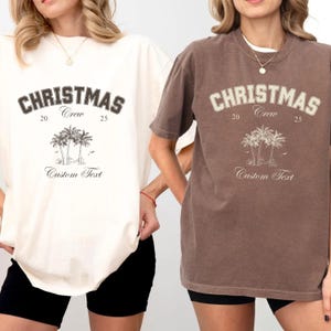 May include: Two short-sleeve t-shirts, one cream and one brown, each featuring the text CHRISTMAS Crew 20 25, a graphic of three palm trees, and the words Custom Text. The shirts have a crew neckline and a relaxed fit design.
