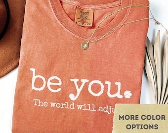 May include: A yam-colored Comfort Colors t-shirt with the words "be you." and "The world will adjust" printed in white. A gold necklace with a pendant rests on the shirt. The shirt is folded, with a tag visible.