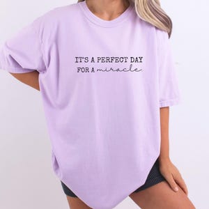 May include: A light purple t-shirt with the text "IT'S A PERFECT DAY FOR A miracle" in black. The shirt has short sleeves and a crew neck.