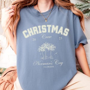 May include: A dusty blue short-sleeve t-shirt featuring a cream-colored graphic with the text CHRISTMAS Crew 20 25, an illustration of three palm trees, and the text Norman's Cay FLORIDA. The shirt has a crew neckline and a relaxed fit.
