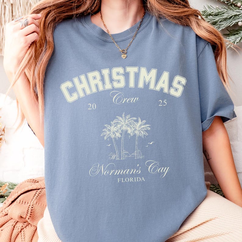 May include: A dusty blue short-sleeve t-shirt featuring a cream-colored graphic with the text CHRISTMAS Crew 20 25, an illustration of three palm trees, and the text Norman's Cay FLORIDA. The shirt has a crew neckline and a relaxed fit.