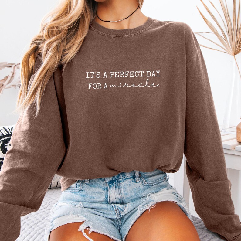 May include: A brown sweatshirt with the text "IT'S A PERFECT DAY FOR A miracle" in white. The sweatshirt has long sleeves and a crew neck. The model is wearing denim shorts.