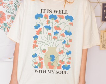 May include: Cream-colored t-shirt featuring a floral design in a vase. The design includes blue and coral flowers with green stems. The text "IT IS WELL WITH MY SOUL" is printed above and below the floral arrangement.