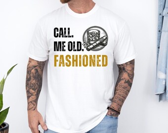 May include: White t-shirt with the text "CALL ME OLD. FASHIONED" in black and gold lettering. A graphic of a cocktail, ice, and a cigar is printed on the shirt. The shirt is a casual style.