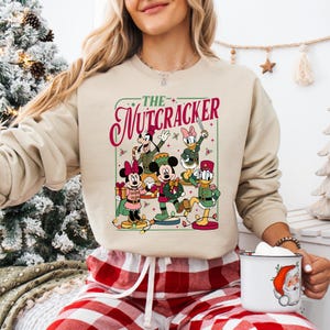 May include: Beige sweatshirt featuring a colorful graphic of Disney characters in Nutcracker costumes. The text "The Nutcracker" is displayed in a decorative font. The sweatshirt is paired with red and white plaid pajama pants.