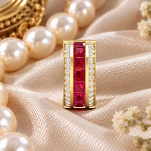 May include: A rectangular gold pendant featuring a central vertical row of five square-cut deep red rubies. The rubies are flanked on both sides by two narrow channels set with small, round, clear white diamonds, creating a structured design.