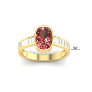 Puede incluir: Un anillo de oro con una gran piedra preciosa de color rosa rojizo de corte ovalado en el centro. La banda también es dorada, con piedras preciosas rectangulares y transparentes a cada lado de la piedra central. Sobre fondo blanco.
