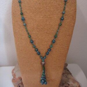 May include: A beaded necklace with a mix of teal and green beads. The necklace has a long, dangling tassel of beads and is displayed on a brown mannequin. The beads vary in size and are strung on a thin, green cord.