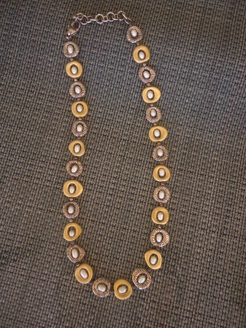 K&ouml;nnte beinhalten: Eine silber- und goldfarbene Halskette mit ovalen und kreisf&ouml;rmigen Gliedern. Die Halskette hat einen Kettenverschluss und abwechselnd goldfarbene ovale Glieder mit silberfarbenen kreisf&ouml;rmigen Gliedern. Jedes Glied hat eine kleine, wei&szlig;e, perlenartige Perle in der Mitte.