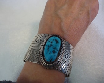 May include: A wide silver cuff bracelet featuring a large, oval-shaped turquoise stone with dark matrix patterns. The silver band is decorated with fine, radiating etched lines and a textured border surrounding the central stone setting.