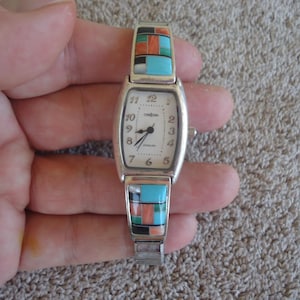 May include: A silver watch with a white face and black Roman numeral hour markers. The watch band is made of silver and has inlaid turquoise, coral, and black stones.