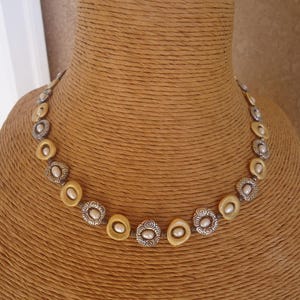 K&ouml;nnte beinhalten: Eine Halskette mit abwechselnd goldfarbenen und silberfarbenen kreisf&ouml;rmigen Gliedern. Die goldenen Glieder haben eine perlenartige Perle in der Mitte. Die silbernen Glieder haben ein dekoratives Wirbelmuster um eine perlenartige Perle in der Mitte. Die Halskette wird auf einer B&uuml;ste pr&auml;sentiert.