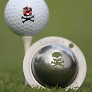 Puede incluir: Pelota de golf blanca con un diseño de calavera y huesos cruzados, sobre un tee. También es visible un marcador de pelota de golf plateado con un recorte de calavera y huesos cruzados. El fondo es hierba verde.