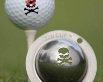 Può includere: Pallina da golf bianca con un disegno a teschio e ossa incrociate, su un tee. È visibile anche un segnaposto per pallina da golf argentato con un ritaglio a teschio e ossa incrociate. Lo sfondo è erba verde.
