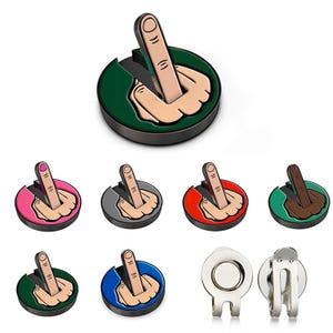 Puede incluir: Colección de marcadores de bola de golf con una mano haciendo el gesto de la peineta. Los marcadores tienen una base redonda en varios colores, incluyendo verde, rosa, rojo y azul. La mano muestra el dedo corazón levantado.