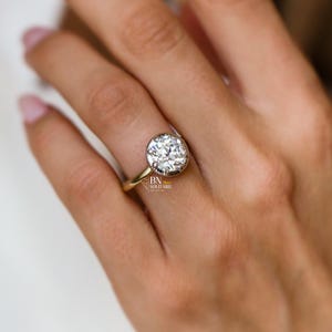 Puede incluir: Un anillo de oro con una piedra preciosa blanca redonda de talla brillante en un engaste de bisel. La banda de metal es fina y pulida. El texto BN SOLITAIRE es visible en letra pequeña en el lateral del engaste, cerca de la base.