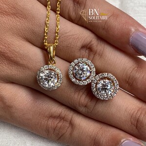 May include: A gold-coloured jewellery set comprising a pendant necklace and matching stud earrings. Each piece features a central, clear gemstone encircled by smaller, clear stones. The necklace chain and earring settings are gold-coloured. The brand name "BN Solitaire" is visible.