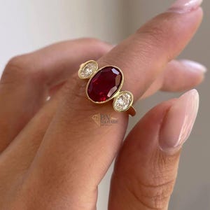Könnte beinhalten: Ein goldener Ring mit einem großen ovalen Rubin-Mittelstein, flankiert von zwei kleineren, runden, klaren Edelsteinen. Der Ring ist in einem goldenen Band gefasst und wird an einem Finger getragen.
