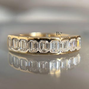 May include: A gold band ring featuring a series of rectangular-cut, clear gemstones. The ring's setting is a warm gold color, and the gemstones are set closely together, creating a continuous line of sparkle. The ring is displayed on a reflective surface.