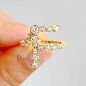 Puede incluir: Un anillo dorado con múltiples gemas redondas y transparentes. El anillo tiene un diseño único, con gemas dispuestas en un patrón curvo y lineal. El anillo está sostenido por una mano. El logotipo "BN SOLITAIRE NEW YORK, NY" es visible.