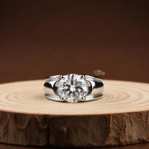Può includere: Un anello color argento con una grande gemma trasparente a taglio brillante rotondo, fissata da quattro griffe. La fascia è larga e liscia con una finitura metallica lucida. Il testo BN SOLITAIRE è visibile vicino alla gemma.