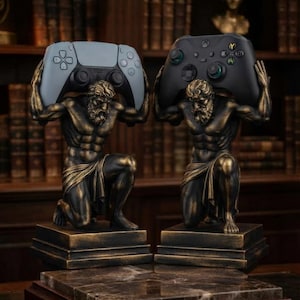 May include: Two bronze-colored statues of kneeling figures with muscular builds, each resting on a square base. One figure holds a light gray gaming controller above its head, while the other figure holds a black gaming controller above its head.