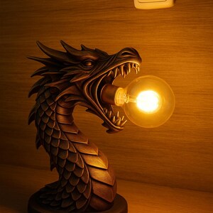 以下が含まれることがあります： ブロンズ色のドラゴンの形をしたランプ。口を開けて丸い電球を咥えている。ランプには詳細な鱗があり、丸い台座に置かれている。背景は木製の壁で、スイッチがある。