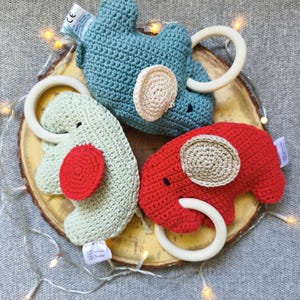 Può includere: Tre giocattoli per bambini a forma di elefante all'uncinetto in blu, verde e rosso. Ogni giocattolo ha un anello di legno e un orecchio di colore a contrasto. I giocattoli sono disposti su una fetta di legno, con uno sfondo grigio e luci.