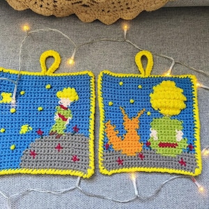 Könnte beinhalten: Zwei quadratische, gehäkelte Topflappen mit gelbem Rand und Aufhängeschlaufen. Jeder zeigt eine Szene aus Der kleine Prinz, mit blauem Himmel, Sternen und einer Figur oder einem Fuchs. Die Designs sind in Gelb, Grün, Orange und Rot gehalten.