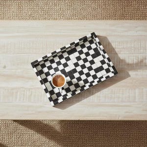 May include: A rectangular serving tray with a black and white checkered pattern. A white cup of coffee sits in the center of the tray. The tray is on a light-colored wooden table.