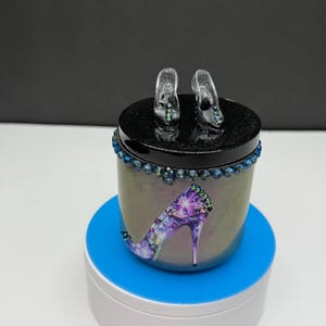 May include: A decorative jar with a black lid and a design of a high-heeled shoe. The jar is decorated with blue and purple rhinestones. Two miniature clear high heels are on top of the lid.