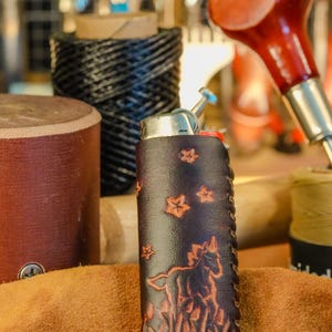 May include: A dark brown leather lighter case with a horse and star design. The case is hand-stitched and features a detailed embossed image of a horse in a field. Other crafting tools are in the background.