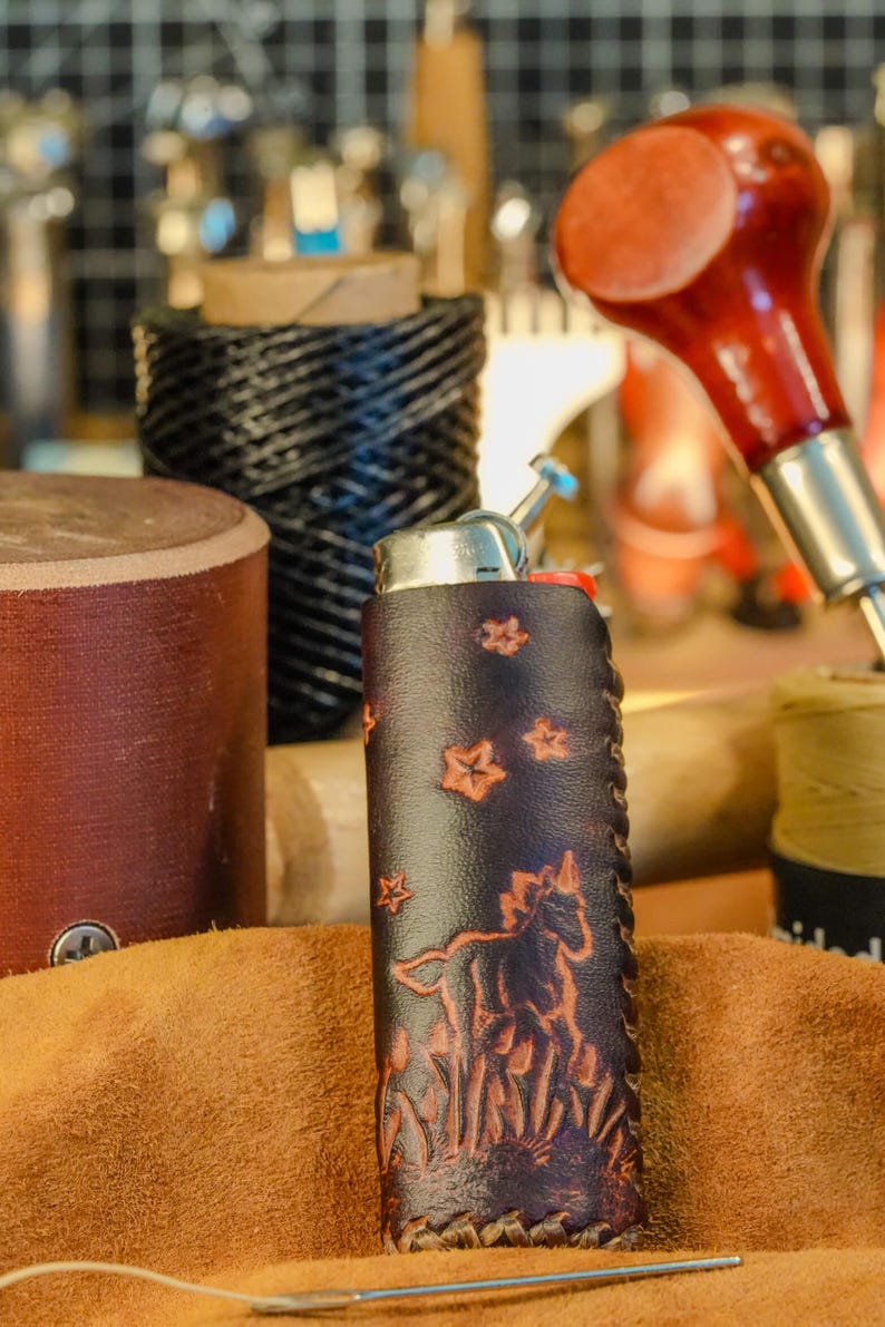 May include: A dark brown leather lighter case with a horse and star design. The case is hand-stitched and features a detailed embossed image of a horse in a field. Other crafting tools are in the background.