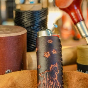 May include: A dark brown leather lighter case with a horse and star design. The case is hand-stitched and features a detailed embossed image of a horse and stars. The lighter case is on a brown surface.