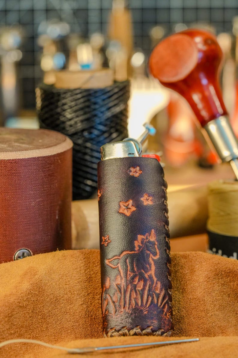 May include: A dark brown leather lighter case with a horse and star design. The case is hand-stitched and features a detailed embossed image of a horse and stars. The lighter case is on a brown surface.