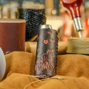 May include: A handcrafted leather flask with a horse and floral design. The dark brown flask is surrounded by leather crafting tools and materials, including spools of thread and a leather hide. The flask is approximately 5 inches tall.