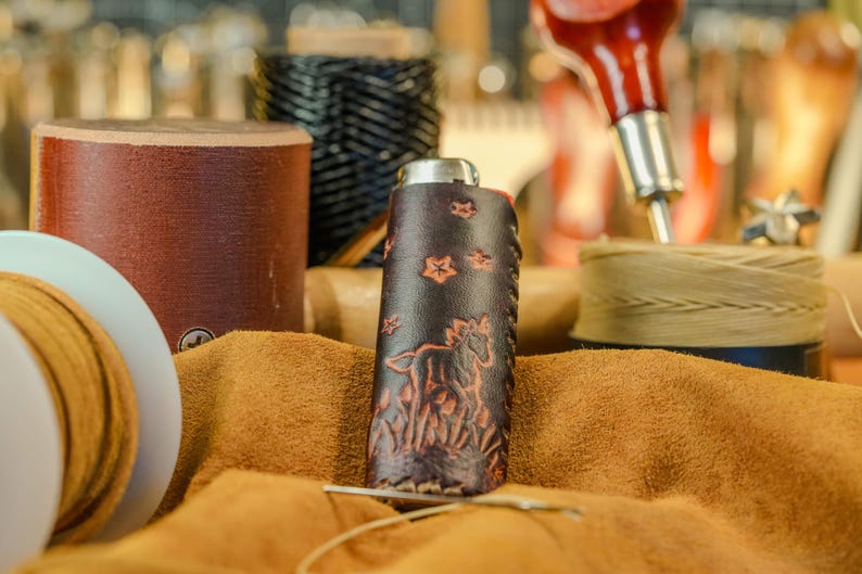 May include: A handcrafted leather flask with a horse and floral design. The dark brown flask is surrounded by leather crafting tools and materials, including spools of thread and a leather hide. The flask is approximately 5 inches tall.