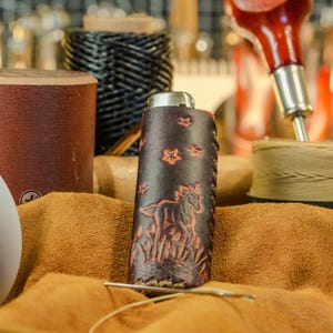 May include: A handcrafted leather flask with a horse and floral design. The dark brown flask is decorated with an embossed horse and flowers. The flask is surrounded by leather crafting tools and materials, including spools of thread and leather.