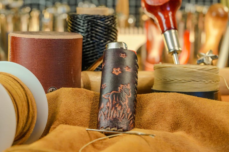 May include: A handcrafted leather flask with a horse and floral design. The dark brown flask is decorated with an embossed horse and flowers. The flask is surrounded by leather crafting tools and materials, including spools of thread and leather.