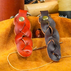 May include: Two braided leather keychains, one red and one dark brown, with gold-colored rivets. The keychains are displayed on a textured orange surface, with a needle and thread visible.