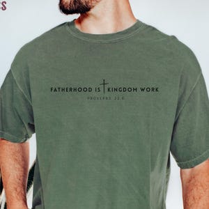 Puede incluir: Camiseta verde musgo con el texto "FATHERHOOD IS | KINGDOM WORK" y "PROVERBIOS 22:6" en negro. La camiseta es de color liso y tiene mangas cortas. La palabra "MOSS" está en la esquina superior izquierda.