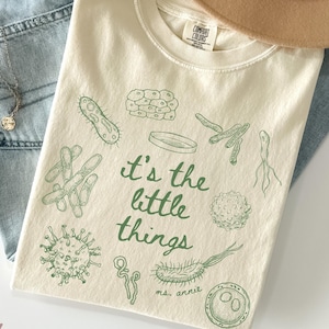 Puede incluir: Una camiseta de manga corta color marfil con un estampado gráfico verde de varios microorganismos y el texto "it's the little things" y "ms. annie". La camiseta tiene cuello redondo y una etiqueta de la marca Comfort Colors en el cuello.