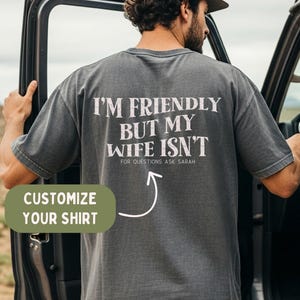 Puede incluir: Una camiseta gris carbón de manga corta con texto blanco desgastado en la espalda que dice I'M FRIENDLY BUT MY WIFE ISN'T y FOR QUESTIONS ASK SARAH junto a una flecha. La camiseta se lleva puesta con una gorra de béisbol de color marrón.