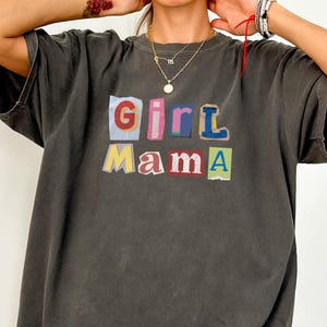 May include: Dark gray t-shirt with the words "Girl Mama" in colorful, cut-out style letters. The shirt has a relaxed fit and short sleeves. The text is in red, blue, yellow, pink, and green.