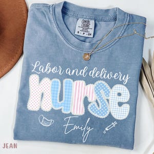 Puede incluir: Camiseta azul vaquero con las palabras "Labor and delivery Nurse" en escritura blanca. La palabra "Nurse" está en un diseño de patchwork con patrones rosas y azules. El nombre "Emily" está debajo de la palabra "Nurse". Un collar dorado está en la camisa.